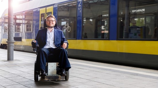 Constantin Grosch hat einen Anzug an und steht auf einem Bahnsteig. Im Hintergrund ist ein blau gelber Zug zu erkennen. Die Sonne reflektiert sich in den Zugscheiben